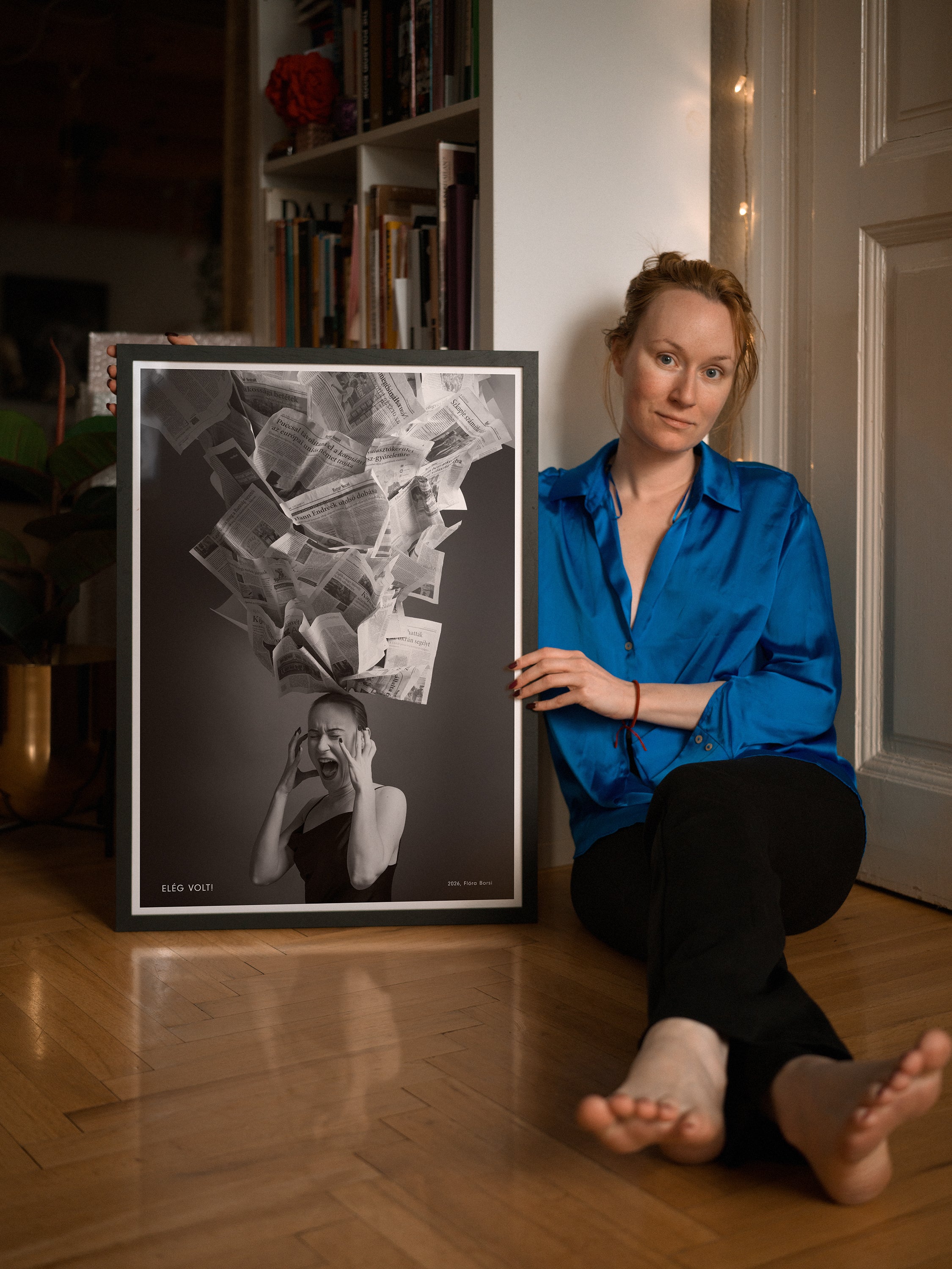 Woman sitting on the floor next to a framed artwork in a room with books and decor.