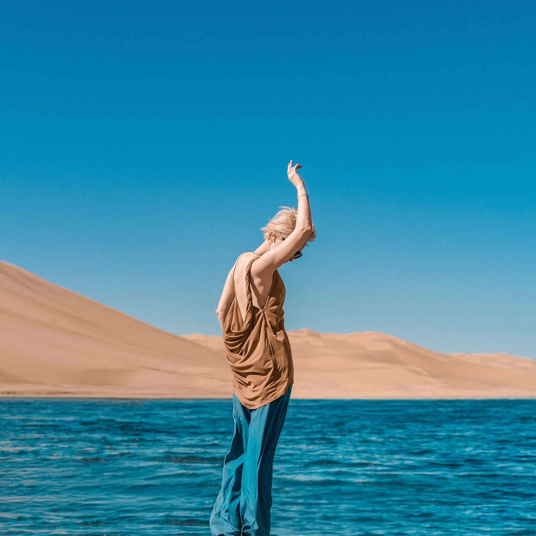 Person standing by a body of water with sand dunes and clear blue sky in the background